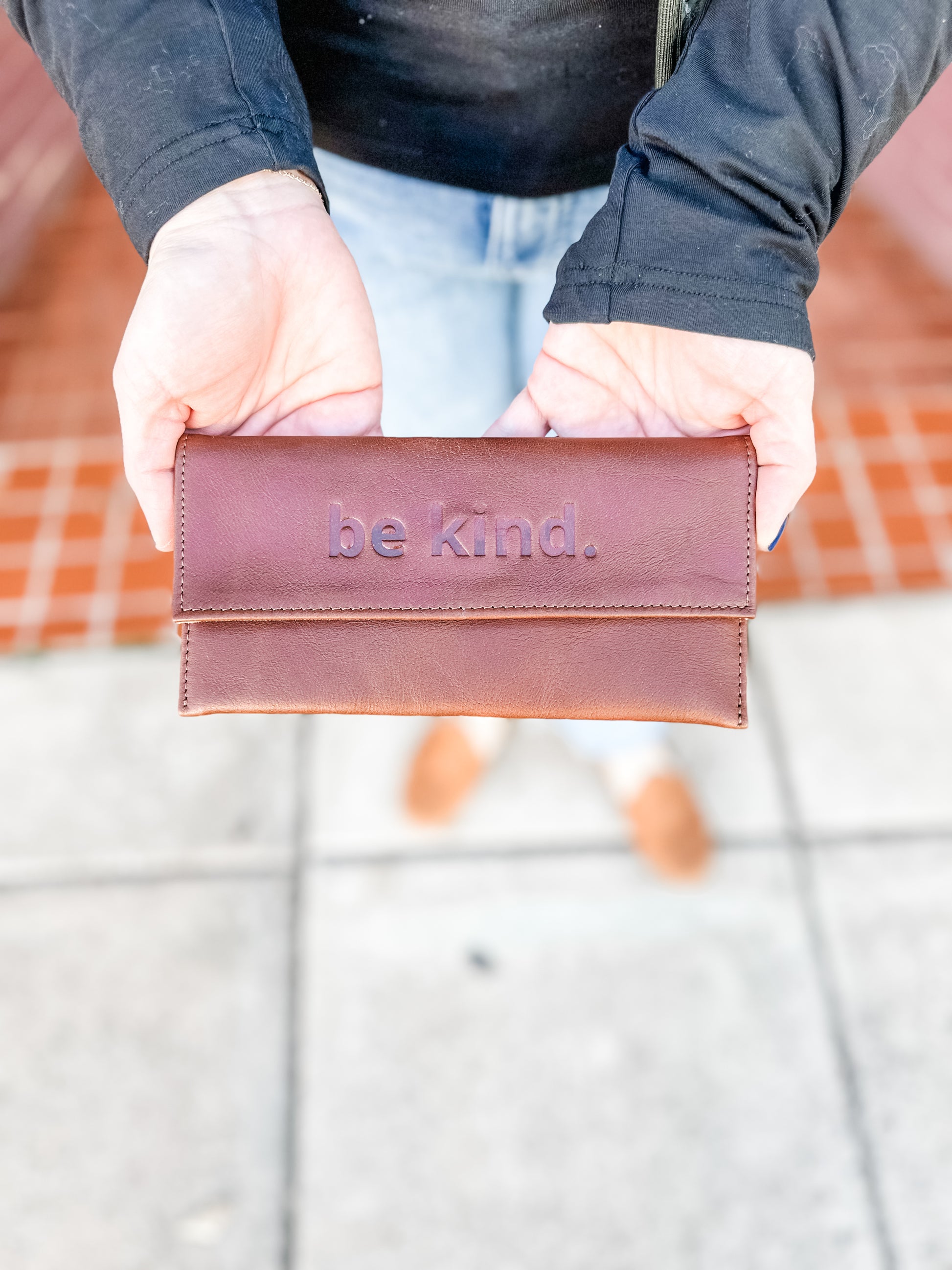 A person holding a brown leather sunglasses case with the text 'be kind' embossed on the front.