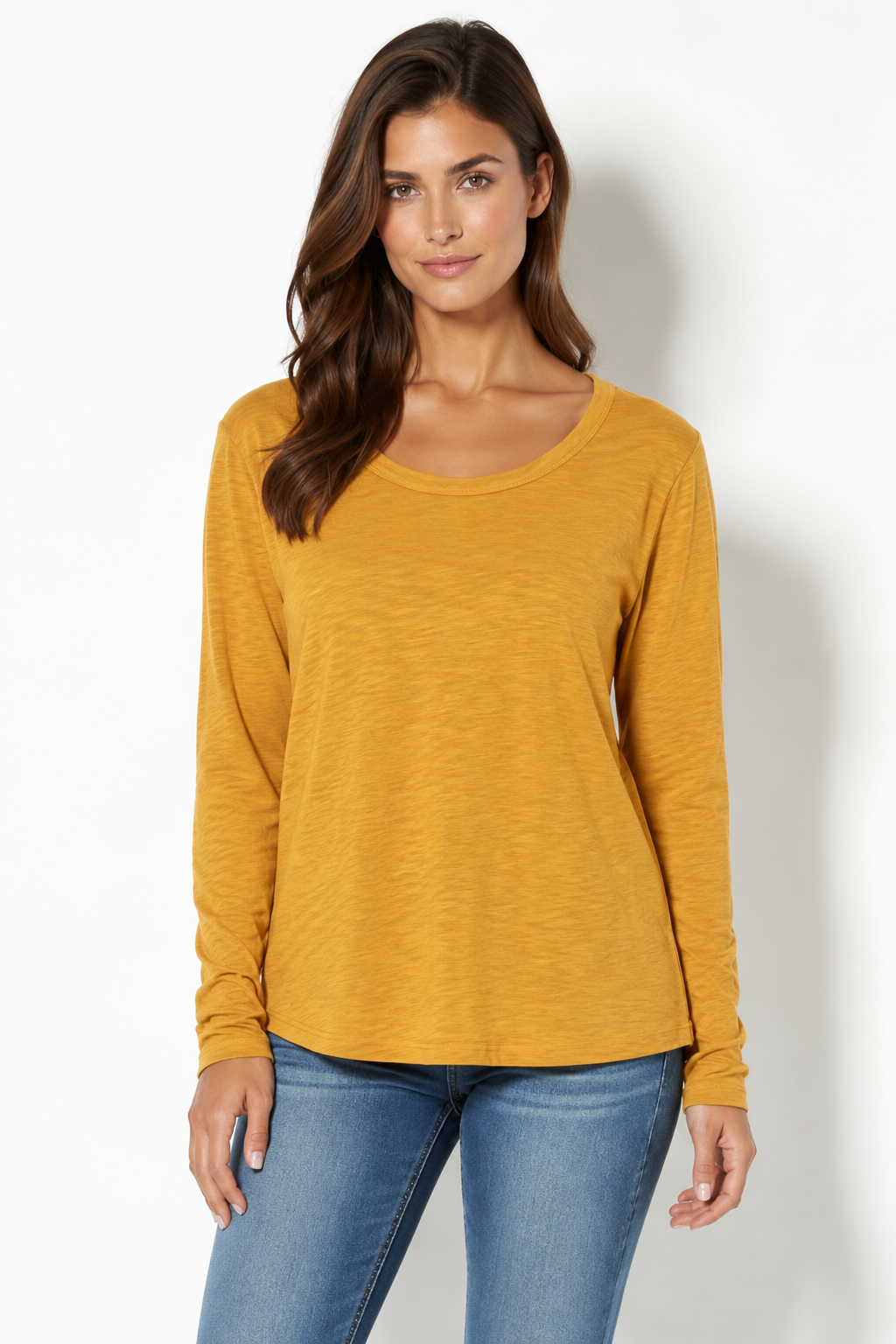 A woman modeling a mustard yellow long-sleeve scoop-neck top paired with blue jeans, standing against a plain white studio background.