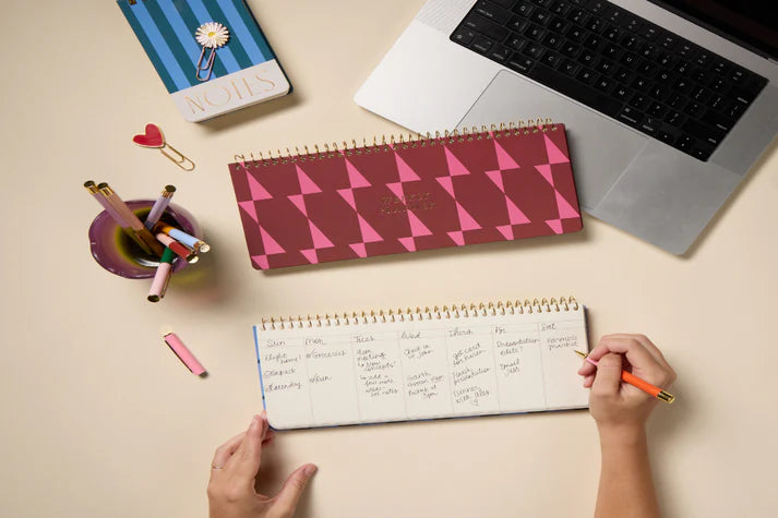 Person writing in a notebook with a pink and red patterned planner and laptop on a desk.