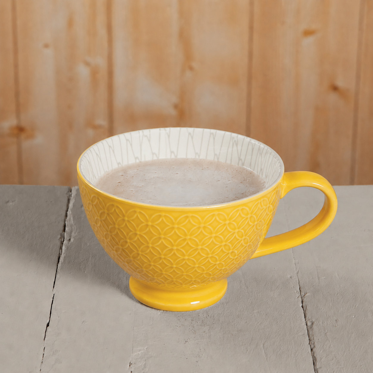 A yellow ceramic mug with a geometric embossed pattern on the exterior and a white interior, filled with a light brown beverage, set on a gray wooden surface against a light wood background.