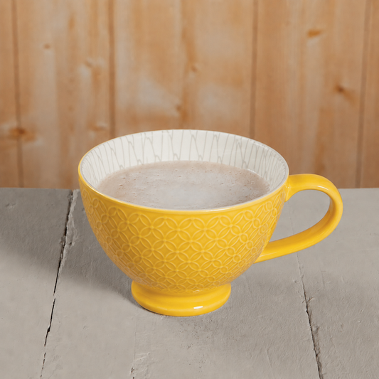A yellow ceramic mug with a geometric embossed pattern on the exterior and a white interior, filled with a light brown beverage, set on a gray wooden surface against a light wood background.